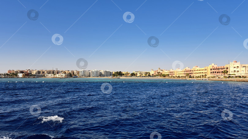 Скачать Вид на побережье с ярко-синими океанскими волнами, разбивающимися о песчаный берег, и разноцветными зданиями, выстроившимися вдоль пляжа под ясным голубым небом. Красное море. Хургада. Египет фотосток Ozero
