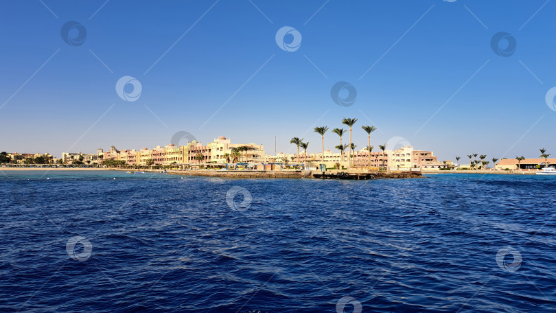 Скачать Прибрежный курорт с пальмами и зданиями вдоль береговой линии, отражающимися в спокойных голубых водах под ясным небом, создает безмятежную атмосферу отдыха. Красное море. Хургада. Египет фотосток Ozero