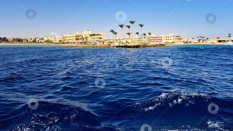 Скачать Живописный вид на побережье с ярко-синими океанскими волнами и далекой береговой линией, усаженной пальмами и зданиями курорта под ясным голубым небом. Красное море. Хургада. Египет фотосток Ozero
