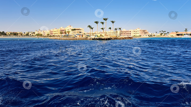 Скачать Живописный прибрежный вид с ярко-синими океанскими волнами и далекой береговой линией с пальмами и зданиями под ясным голубым небом навевает ощущение спокойствия и релаксации. Красное море. Хургада. Египет фотосток Ozero