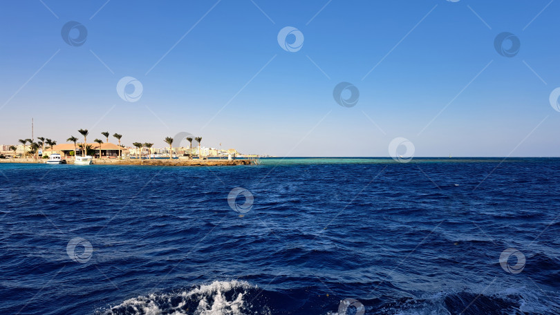 Скачать Яркое побережье с чистыми голубыми водами и отдаленной береговой линией с пальмами и пляжными постройками под ярким солнечным небом. Красное море. Хургада. Египет фотосток Ozero