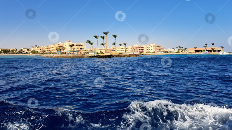 Скачать Живописный вид на побережье с ярко-синими океанскими волнами, пальмами и зданиями курорта под ясным небом создает спокойную морскую атмосферу. Красное море. Хургада. Египет фотосток Ozero