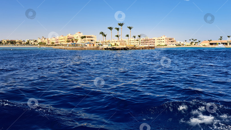 Скачать Яркий прибрежный пейзаж со спокойными синими океанскими волнами, мягко покачивающимися пальмами и отдаленным видом курорта под ясным голубым небом создает атмосферу безмятежного отдыха. Красное море. Хургада. Египет фотосток Ozero