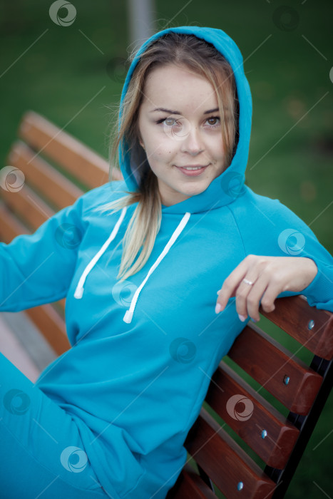 Скачать Улыбающаяся девушка в синем спортивном костюме сидит на деревянной скамейке на открытом воздухе и смотрит в камеру фотосток Ozero