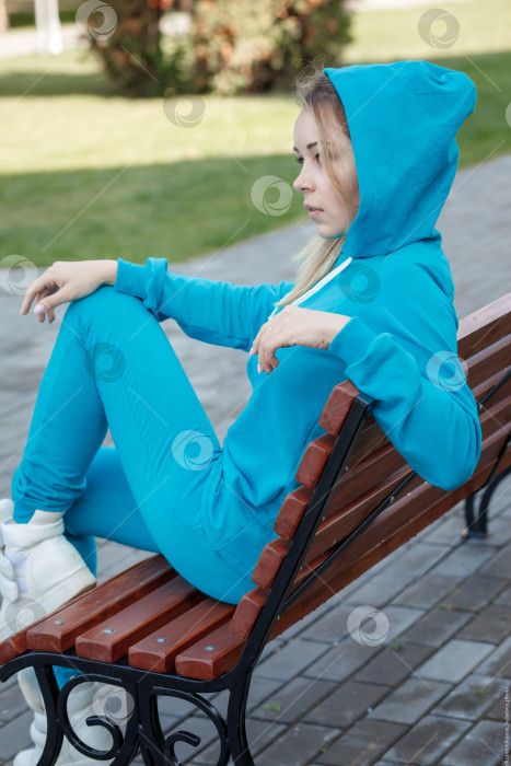 Скачать Улыбающаяся девушка в синем спортивном костюме сидит на деревянной скамейке на открытом воздухе и смотрит в камеру фотосток Ozero