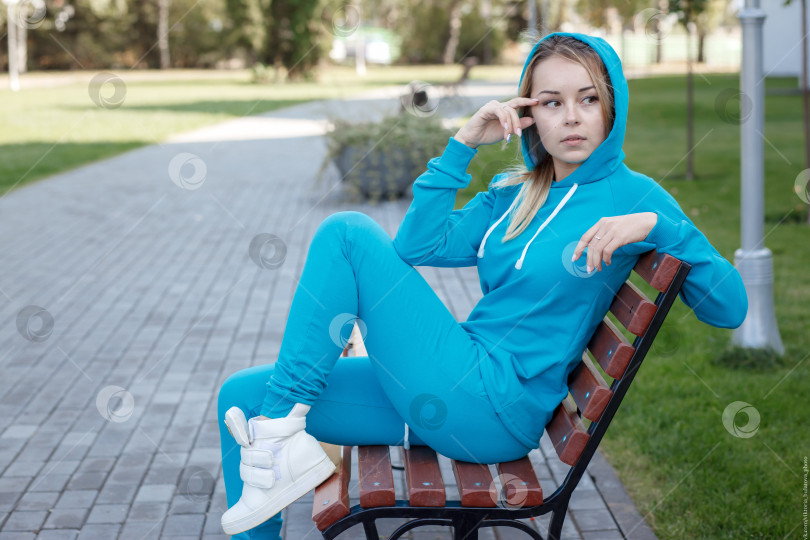 Скачать Улыбающаяся девушка в синем спортивном костюме сидит на деревянной скамейке на открытом воздухе и смотрит в камеру фотосток Ozero