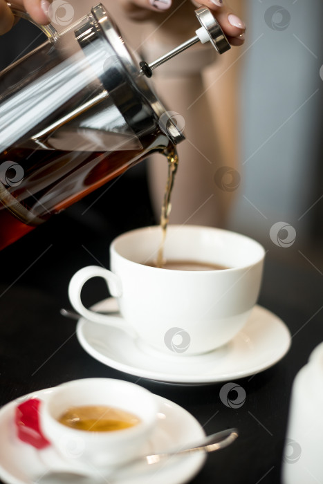 Скачать Рука наливает горячий чай из стеклянного чайника в белую чашку, рядом с чашкой лежит слоеная выпечка на маленькой тарелочке. Обстановка в помещении, естественное освещение из окна. Снимок крупным планом. фотосток Ozero