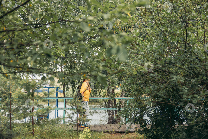 Скачать мужчина в оранжевой толстовке с капюшоном, стоящий на небольшом деревянном мосту с рюкзаком, пышная зеленая листва и деревья у воды, экологический туризм, туристическое агентство, компания по организации активного отдыха фотосток Ozero