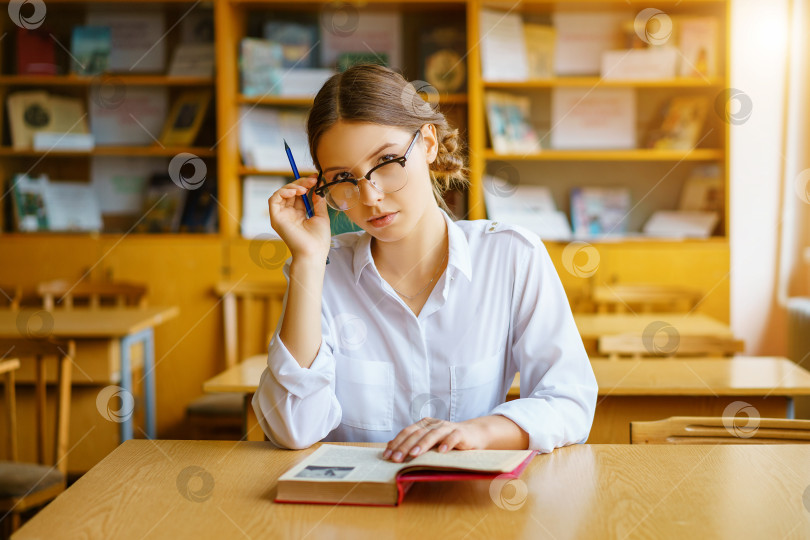 Скачать девушка в очках сидит за столом с книгой в классе, держит карандаш в руке, задумчивый вид. фотосток Ozero