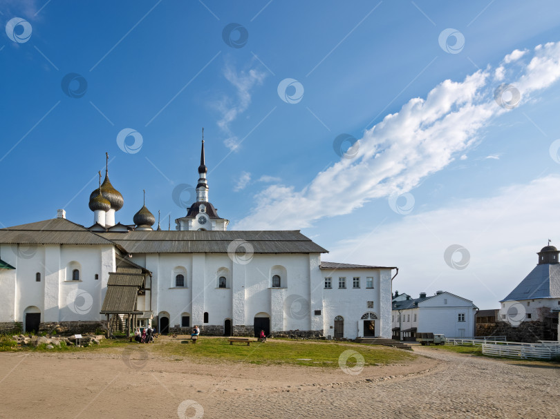 Скачать Площадь перед Трапезным храмом в Соловецком монастыре, вымощенная камнем и грунтом, залита летним солнечным светом. На заднем плане возвышаются белые стены монастырского комплекса с характерными куполами и высокой колокольней, уходящими в ясное голубое небо с легкими облаками фотосток Ozero