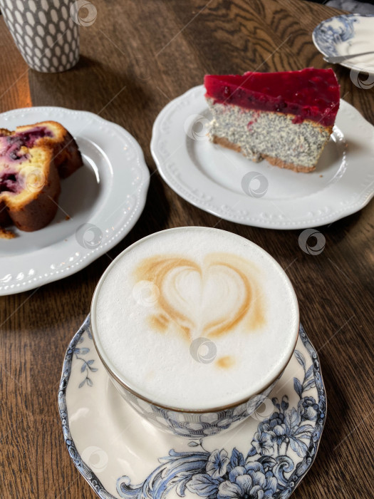 Скачать Кофейный латте-арт с сердечками рядом с двумя тарелками пирожных на деревянном столе фотосток Ozero