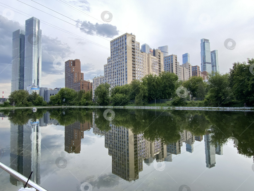 Скачать Городской пейзаж отражается в спокойной воде озера, окруженного пышными зелеными деревьями и облачным небом фотосток Ozero