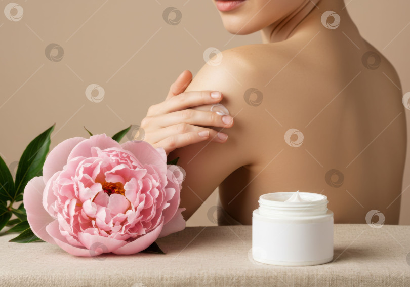 Скачать Женщина, наносящая крем для тела на плечо, рядом с ней баночка с кремом и цветок пиона. Процедура по уходу за кожей. фотосток Ozero