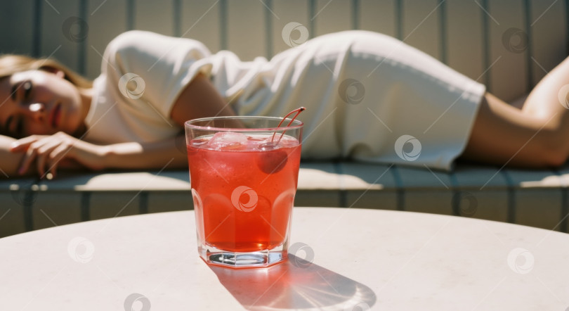 Скачать Красный коктейль со льдом и вишней на столе с размытой женщиной, отдыхающей на заднем плане, для летнего напитка или расслабляющего отдыха. фотосток Ozero