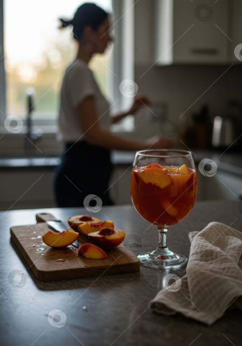 Скачать Освежающий домашний персиковый напиток в бокале с нарезанными персиками на доске, на заднем плане изображена женщина. фотосток Ozero