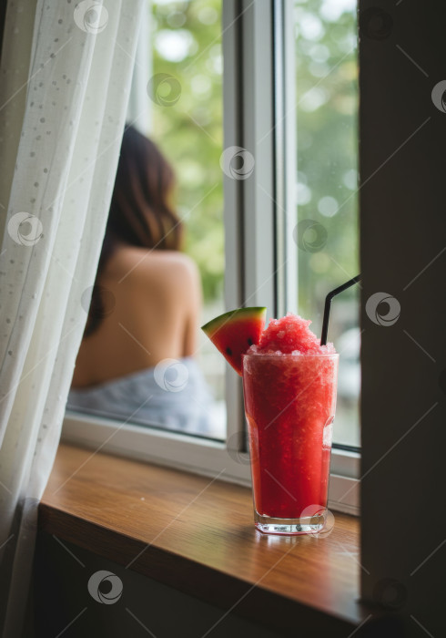 Скачать Холодный освежающий арбузный смузи с соломинкой и ломтиком фрукта на деревянном подоконнике и женщиной на заднем плане. Напиток для летнего отдыха. фотосток Ozero