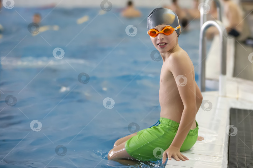 Скачать Мальчик в плавательной шапочке и защитных очках сидит на бортике спортивного бассейна. фотосток Ozero