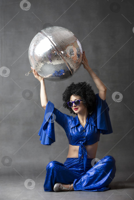 Скачать Женщина в расклешенном костюме семидесятых годов с дискотечным шаром в парике-афро на сером фоне. фотосток Ozero
