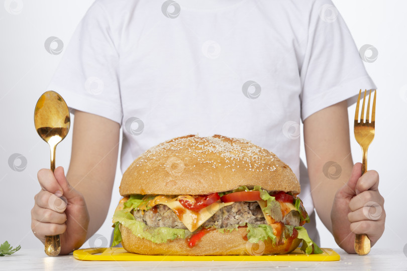 Скачать Свежий вкусный бургер на фоне мужчины, который держит в руках золотую вилку и ложку. Элитный фаст-фуд. Роскошный гамбургер. фотосток Ozero