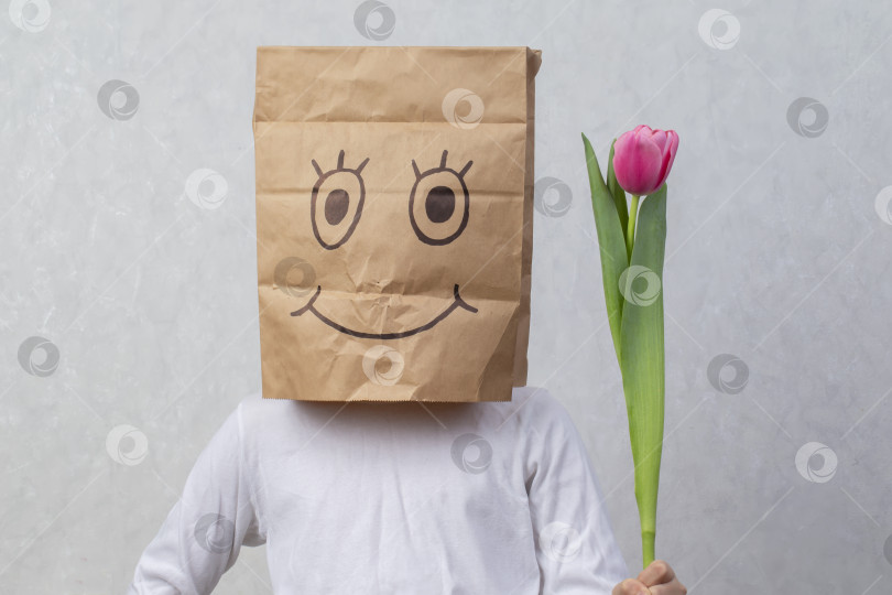 Скачать Ребенок с картонной коробкой на голове, на которой маркером нарисована улыбка, держит в руках тюльпан. Концепция покупки праздничных подарков. фотосток Ozero