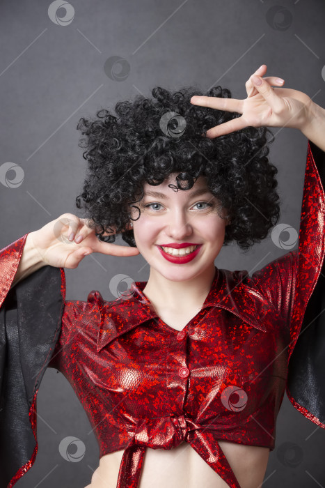 Скачать Красивая девушка в красном дискотечном костюме и черном кудрявом парике улыбается. фотосток Ozero