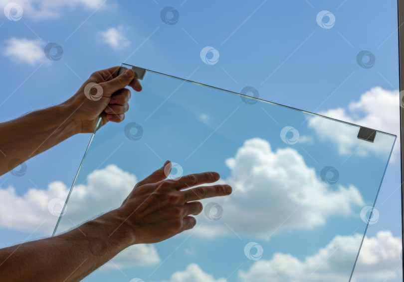 Скачать Мужчина поднимает прозрачное стекло к голубому небу с облаками. Строительные материалы, установка окон и концепция остекления. фотосток Ozero