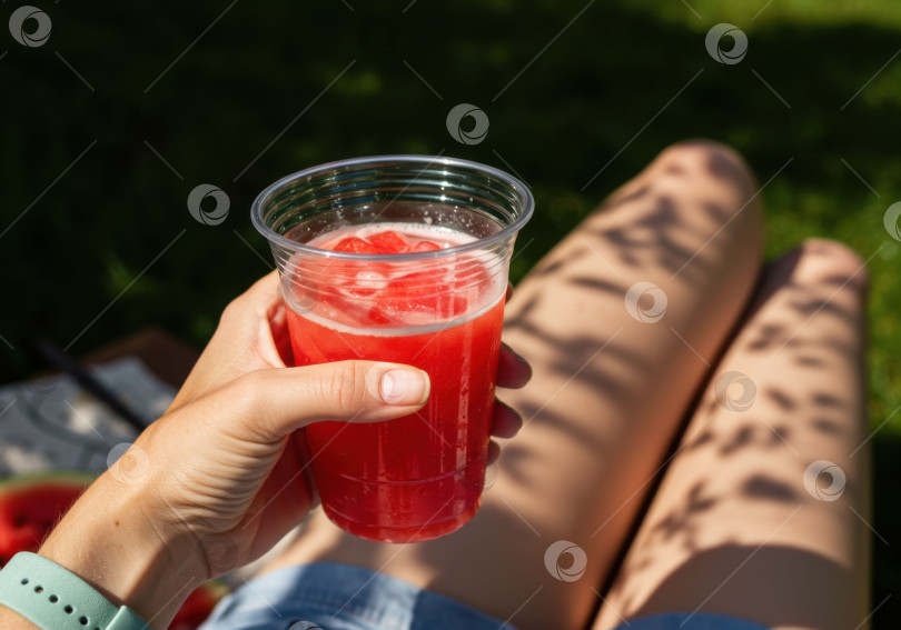 Скачать Женская рука держит красный холодный напиток со льдом в пластиковом стаканчике, отдыхая на свежем воздухе солнечным летним днем. Концепция освежающего напитка. фотосток Ozero