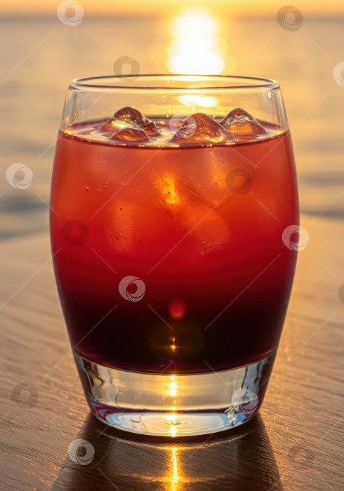 Скачать Крупный план красного коктейля со льдом на фоне заката над водой, отражающего теплый свет на столе. фотосток Ozero