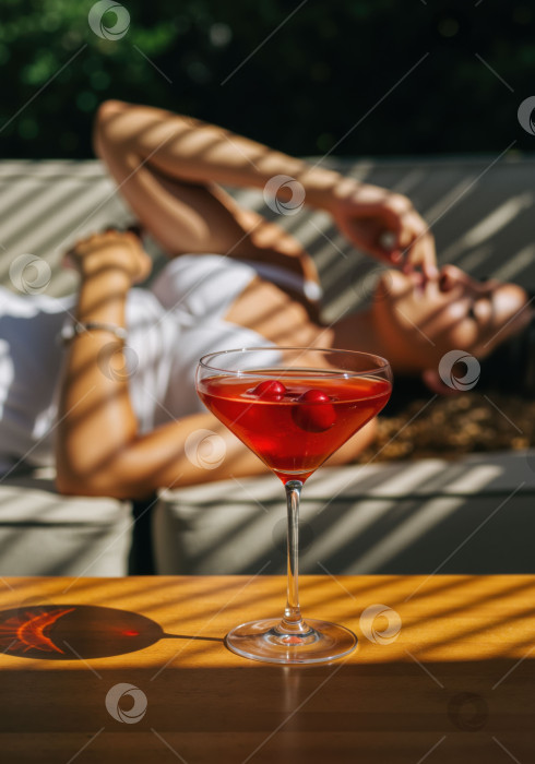 Скачать Красный коктейль с вишнями на столе на фоне размытой женщины, развалившейся на заднем плане, - напиток для летнего отдыха. фотосток Ozero