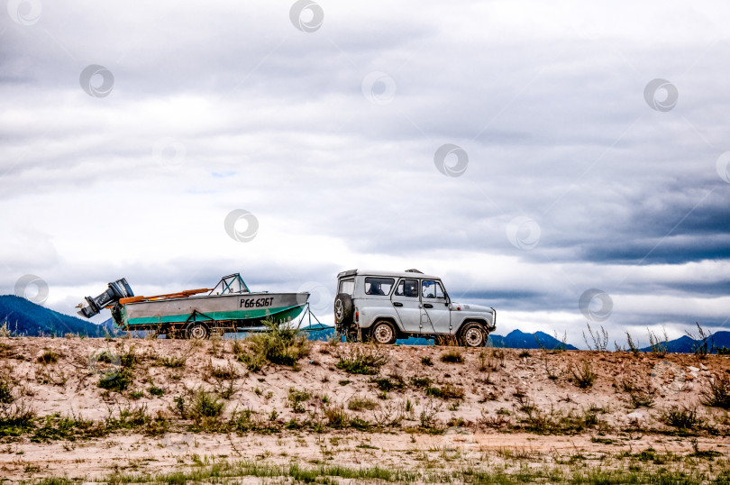 Скачать Байкал, Россия, 2 мая 2018 года, внедорожник с рыбацкой лодкой в прицепе, летний пейзаж в пасмурный день. фотосток Ozero