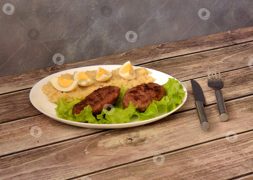 Скачать Тарелка с вкусным обедом, две жареные котлеты на листьях салата и картофельное пюре с нарезанным вкрутую куриным яйцом на деревянном столе, рядом вилка и столовый нож. фотосток Ozero