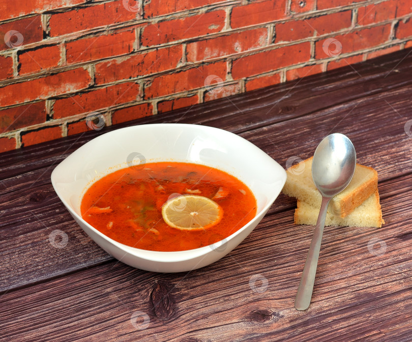 Скачать Солянка, вкусный суп в глубокой миске с ломтиком лимона на деревянном столе, столовой ложкой и двумя кусочками пшеничного хлеба рядом. фотосток Ozero