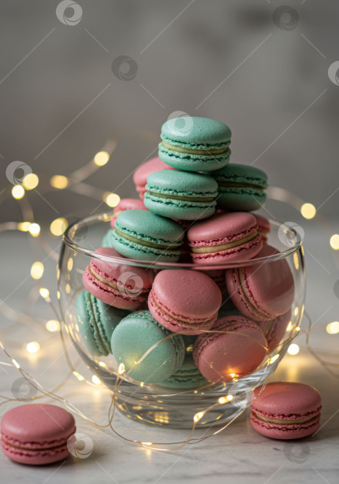 Скачать Горка разноцветных макарон в стеклянной вазочке с гирляндами. Сладкий десерт для праздничного стола и особых случаев. фотосток Ozero
