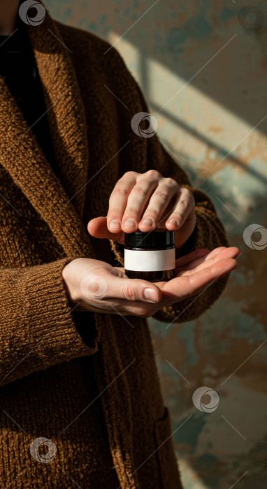Скачать Мужчина в коричневом свитере держит в руках баночку с косметическим средством. Концепция мужского ухода за кожей на коричневом фоне. фотосток Ozero