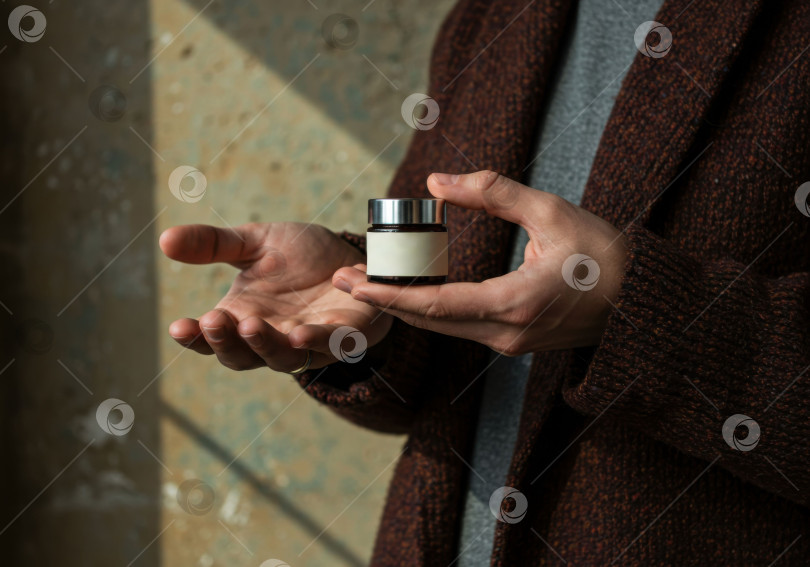 Скачать Мужчина держит в руках коричневую косметическую баночку с пустой этикеткой, демонстрирующую средство для ухода за кожей для мужчин фотосток Ozero