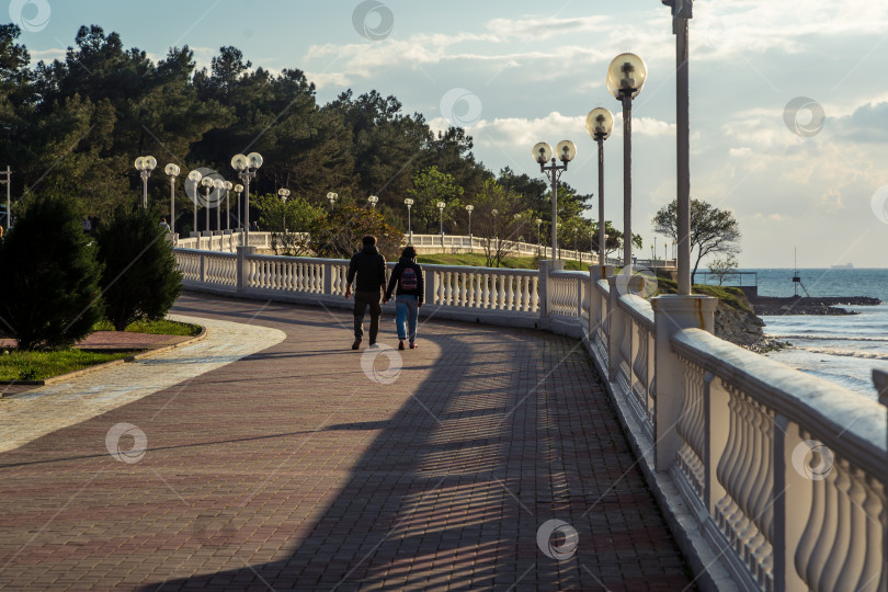 Скачать Город Геленджик. Курортная набережная с балюстрадой и фонарями. Молодая пара прогуливается по пирсу, держась за руки. Солнце садится, и балюстрада отбрасывает тень на набережную. фотосток Ozero