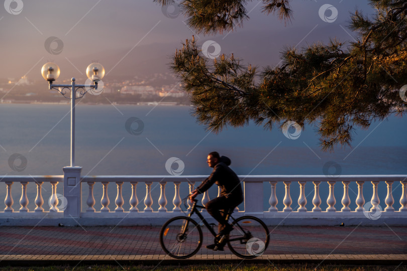 Скачать Курорт Геленджик. Набережная на закате. Балюстрада и набережная в косых лучах заходящего солнца. Велосипедист едет по набережной фотосток Ozero