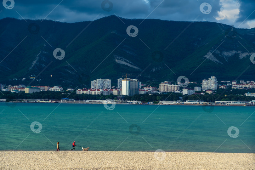 Скачать Курорт Геленджик. Закат. Мужчина и женщина с собакой прогуливаются по пляжу Геленджикской бухты на фоне Кавказских гор. фотосток Ozero