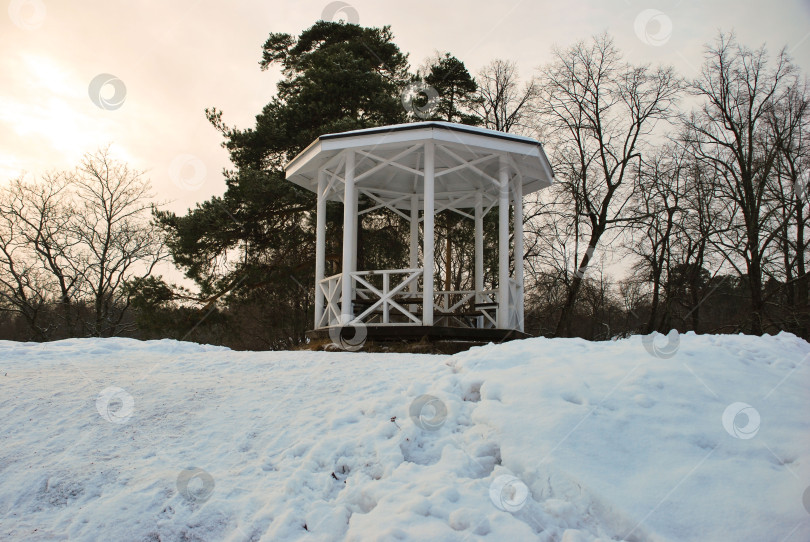 Скачать Белая беседка в заснеженном парковом пейзаже с деревьями и закатным небом фотосток Ozero