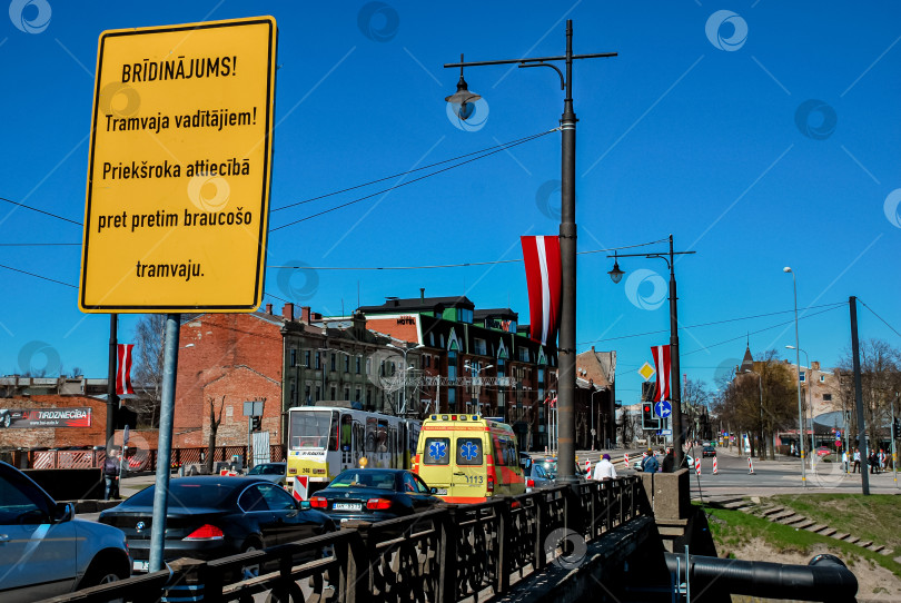 Скачать Предупреждающий знак на городской улице с оживленным движением и латвийскими флагами в Риге в солнечный день фотосток Ozero
