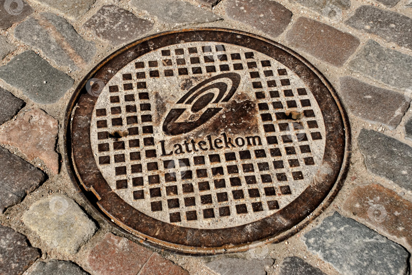 Скачать Крышка канализационного люка Lattelekom на мощеной улице фотосток Ozero