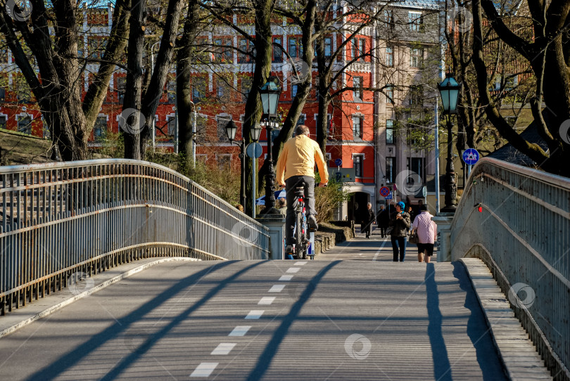 Скачать Взрослый кавказский мужчина едет на велосипеде по городскому пешеходному мосту на фоне деревьев и людей фотосток Ozero
