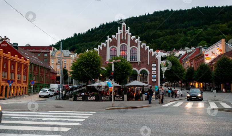 Скачать Живописная городская площадь с историческим кирпичным зданием и пышным зеленым холмом на заднем плане фотосток Ozero