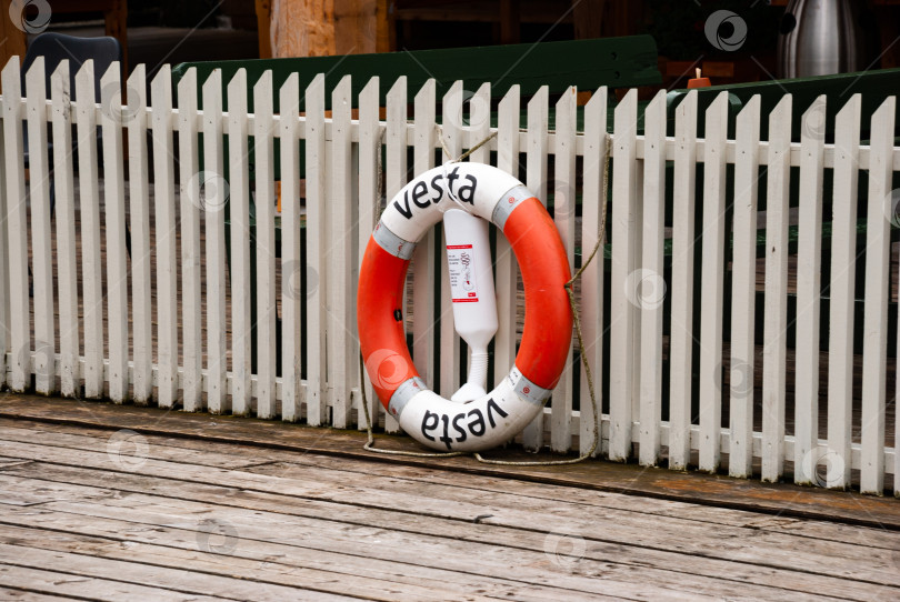 Скачать Бело-оранжевый спасательный круг на деревянном пирсе на фоне белого забора фотосток Ozero