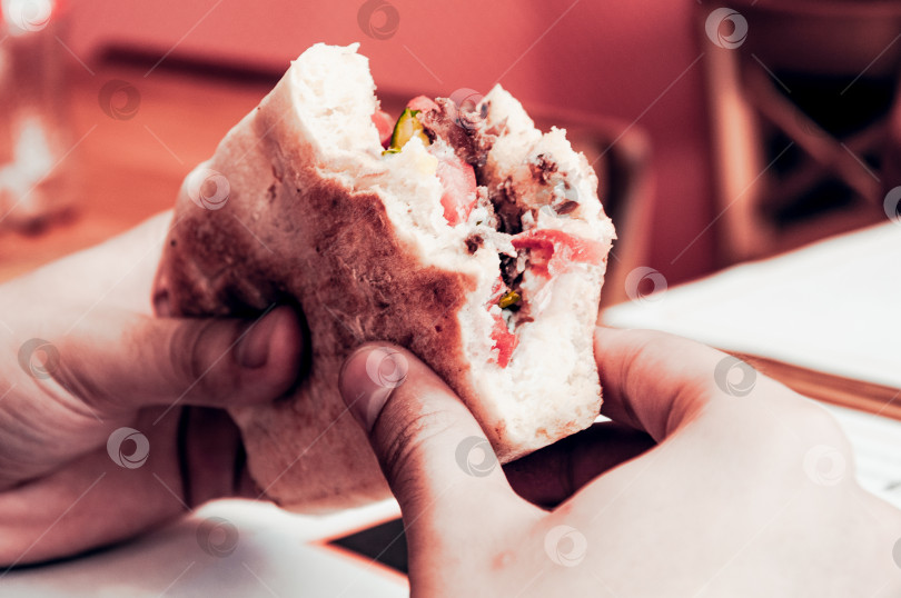 Скачать Надкусанный сэндвич панини с овощами и мясом держит в руках посетитель ресторана фотосток Ozero