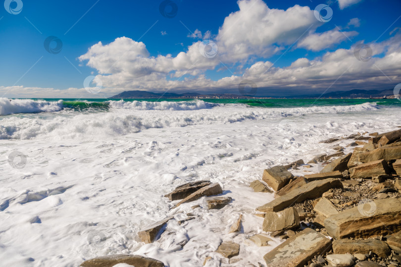 Скачать Курорт Кабардинка. Большие зеленые штормовые волны в белой пене накатываются на каменистый пляж Кабардинки. На заднем плане - порт Новороссийск и Кавказские горы. Большие камни на берегу. Брызги и пена от волн фотосток Ozero