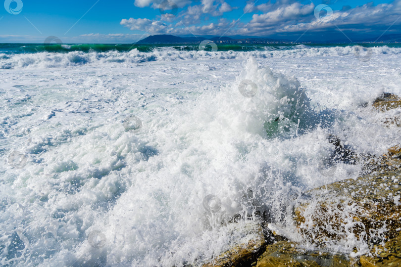 Скачать Курорт Кабардинка. Большие зеленые штормовые волны в белой пене накатываются на каменистый пляж Кабардинки. На заднем плане - порт Новороссийск и Кавказские горы. Большие камни на берегу. Брызги и пена от волн фотосток Ozero