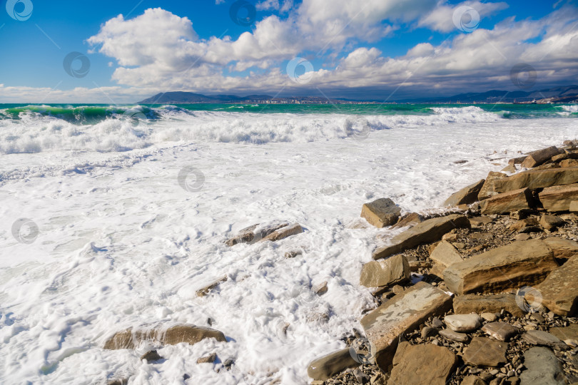 Скачать Курорт Кабардинка. Большие зеленые штормовые волны в белой пене накатываются на каменистый пляж Кабардинки. На заднем плане - порт Новороссийск и Кавказские горы. Большие камни на берегу. Брызги и пена от волн фотосток Ozero