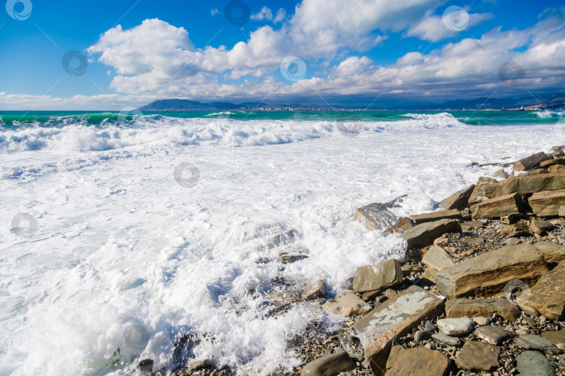 Скачать Курорт Кабардинка. Большие зеленые штормовые волны в белой пене накатываются на каменистый пляж Кабардинки. На заднем плане - порт Новороссийск и Кавказские горы. Большие камни на берегу. Брызги и пена от волн фотосток Ozero
