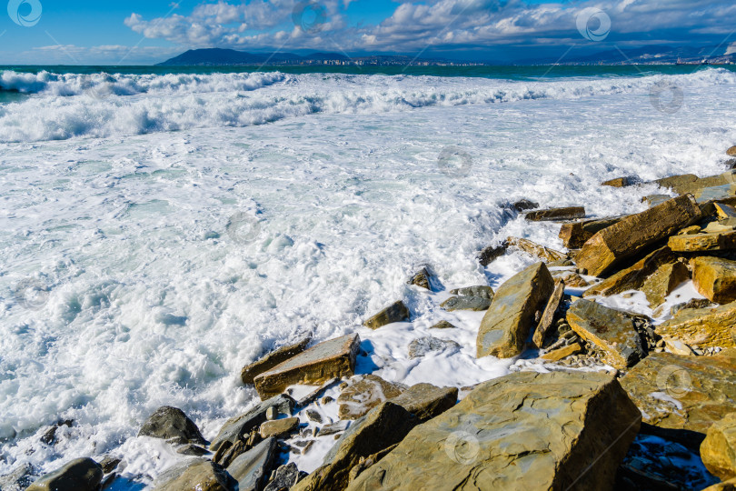 Скачать Курорт Кабардинка. Большие зеленые штормовые волны в белой пене накатываются на каменистый пляж Кабардинки. На заднем плане - порт Новороссийск и Кавказские горы. Большие камни на берегу. Брызги и пена от волн фотосток Ozero